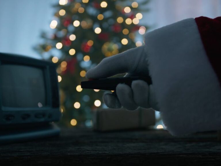 deda mraz uključuje televizor uz pomoć daljinskog upravljača, u pozadini pokloni i jelka